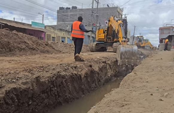 A person with a reflector stand infront of an excavator digging a trench